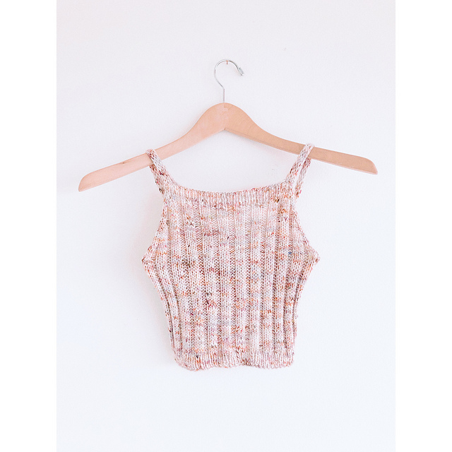 a handknit cropped tank top in a peach color, made with 2 by 2 rib, hanging on a wooden hanger in front of a white background